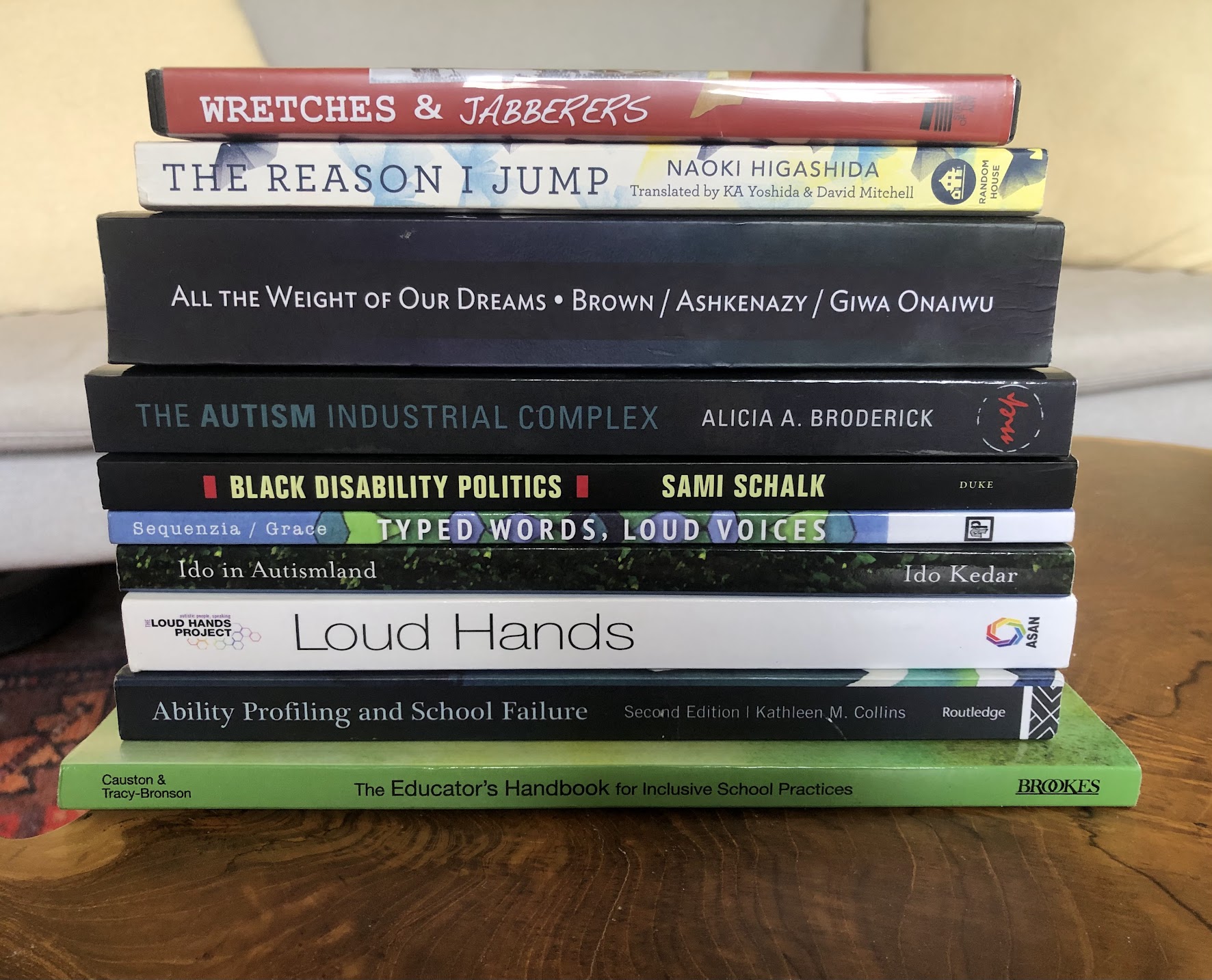 Stack of books and one DVD sitting on a wood table. The titles from top to bottom are: Wretches & Jabberers, The Reason I Jump, All the Weight of Our Dreams, The Autism Industrial Complex, Black Disability Politics, Typed Words, Loud Voices, Ido in Autismland, Loud Hands, Ability Profiling and School Failure, and The Educator's Handbook for Inclusive School Practices.