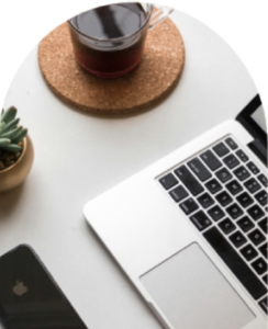 Birds-eye view of the corners of a few objects that sit on a white surface: a silver macbook, a clear glass of tea on a cork coaster, a potted succulent, and a black iphone.