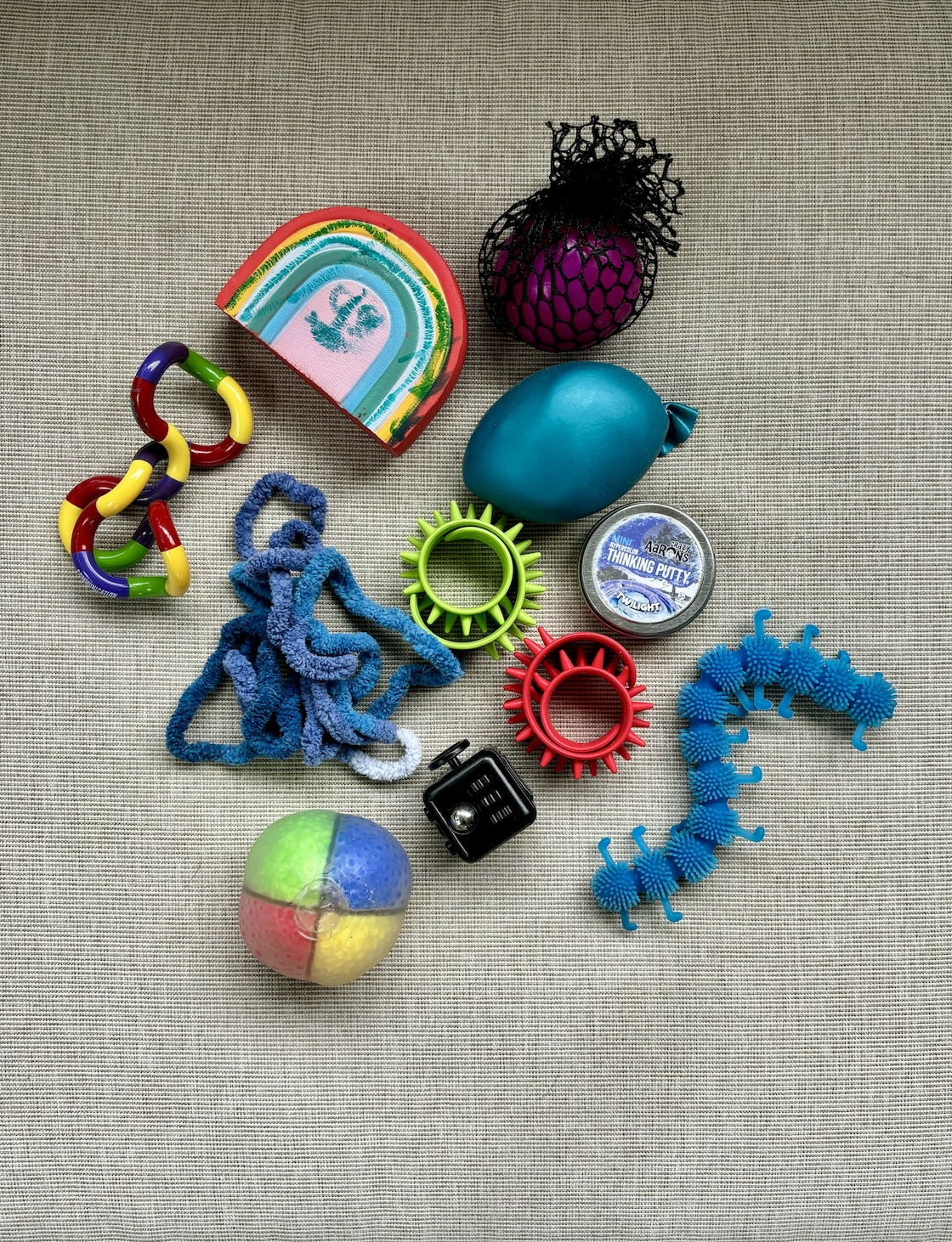 A birds-eye view of an assortment of colorful fidget toys in a pile, laying on a fabric surface. Toys include a clear squishy ball with colored sections of beads inside, a blue fuzzy string in a pile, lime green and red spikey slap bracelets, a black cube with different black and silver dials on each side, a turquoise rubber caterepillar with tiny spikes and missing legs, a round tin labeled Aaron's Thinking Putty, a dense sand ball wrapped in teal rubber, a squishy magenta ball wrapped in black netting, and a squishy rainbow with green marker scribbles from a child.