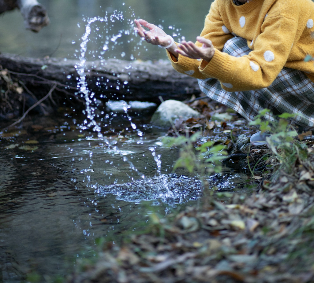 The hands of a white person in a yellow sweater with white polka dots and plaid bottoms are seen splashing water upwards in the woods. Dried leaves sit in the bottom right corner and green plants sprout where the ground meets the water. The person crouches down over the edge.