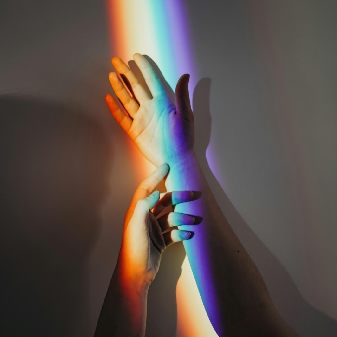 A rainbow ray of light shines down from above onto a hand laying flat with the back of the arm against the wall and another hand gently rests on the other's forearm. The background is dark grey, with the shadow of one hand faintly visible.