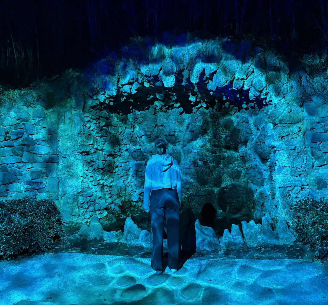 A scene from an event called Garden Glow at Maymont Park. There is a teal light with ranging gradient that shines on the opening of a wall made of stones. The opening has stones that almost look like teeth above and below. We see the back of Liz, standing in front of the opening, staring into it and slightly up.