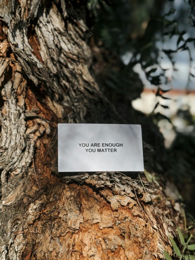 A business-card sized piece of white paper gently rests against and on the bark of a tree. The card says in black text, "You are enough, you matter."